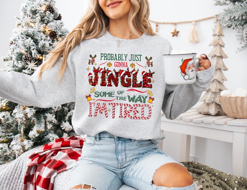 May include: A light gray sweatshirt with the text "Probably Just Gonna Jingle Some of the Way I'm Tired" in red, green, and black. The sweatshirt features Christmas-themed graphics. A white mug with a Santa Claus design is held in the hand.