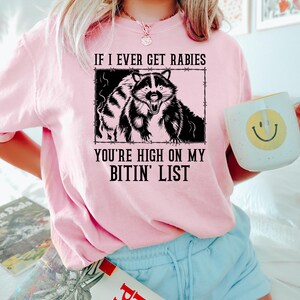 May include: A light pink sweatshirt with the text "IF I EVER GET RABIES YOU'RE HIGH ON MY BITIN' LIST" and a black and white raccoon graphic. The person is holding a white mug with a yellow smiley face.