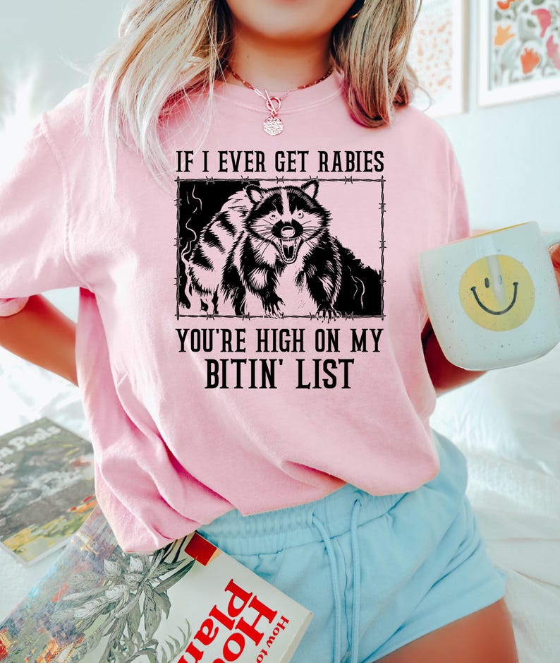 May include: A light pink sweatshirt with the text "IF I EVER GET RABIES YOU'RE HIGH ON MY BITIN' LIST" and a black and white raccoon graphic. The person is holding a white mug with a yellow smiley face.