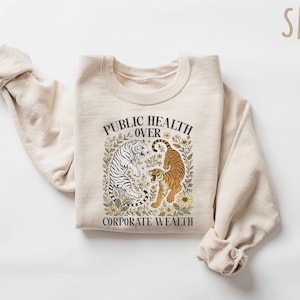 Puede incluir: Sudadera color arena con el texto "PUBLIC HEALTH OVER CORPORATE WEALTH" y un gr&aacute;fico de dos tigres. Uno es blanco con rayas negras, el otro naranja con rayas negras. El dise&ntilde;o est&aacute; rodeado de elementos florales.