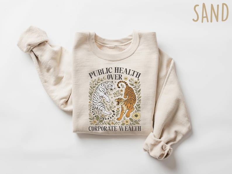 Puede incluir: Sudadera color arena con el texto "PUBLIC HEALTH OVER CORPORATE WEALTH" y un gr&aacute;fico de dos tigres. Uno es blanco con rayas negras, el otro naranja con rayas negras. El dise&ntilde;o est&aacute; rodeado de elementos florales.
