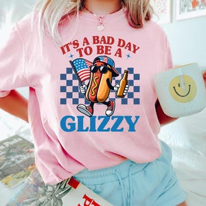 May include: Pink t-shirt with a cartoon hot dog wearing sunglasses and a baseball cap holding a beer and an American flag. The text on the shirt reads "It's a bad day to be a Glizzy".