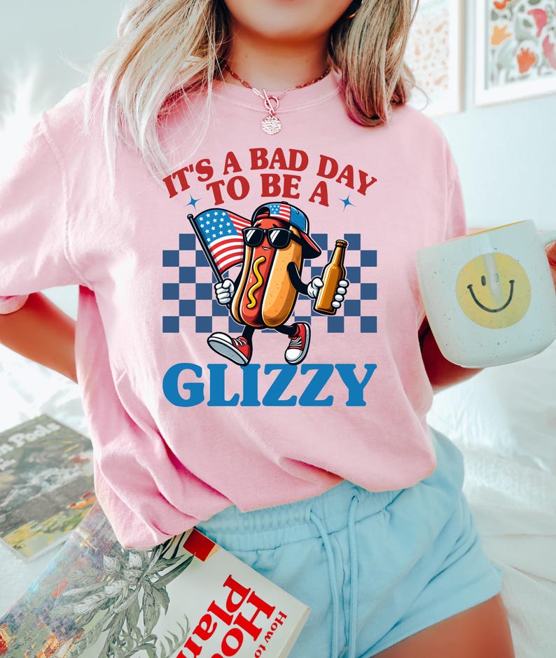 May include: Pink t-shirt with a cartoon hot dog wearing sunglasses and a baseball cap holding a beer and an American flag. The text on the shirt reads "It's a bad day to be a Glizzy".