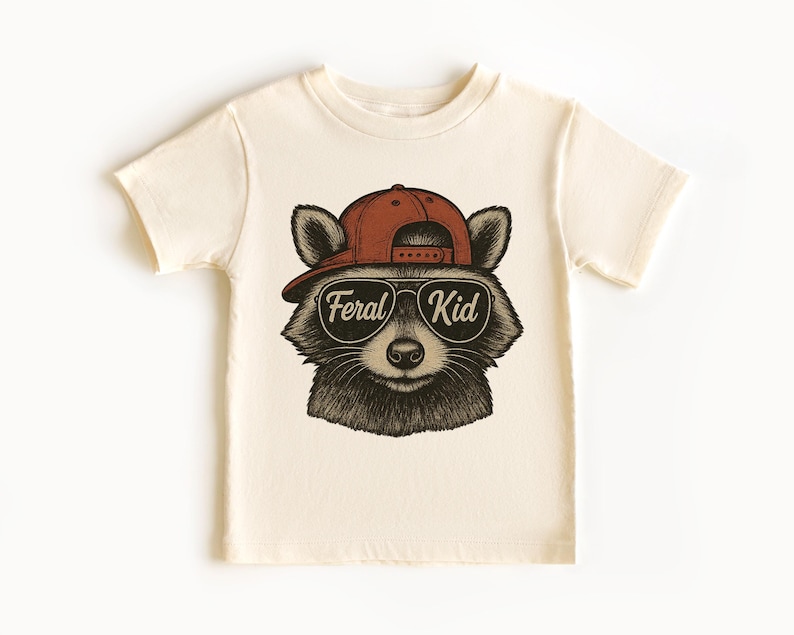 May include: Cream-colored t-shirt featuring a raccoon wearing a red baseball cap and sunglasses. The raccoon graphic has the text "Feral Kid" in a white font. The shirt is a short-sleeved crew neck.