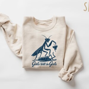 May include: Sand-colored sweatshirt with a navy blue graphic of a praying mantis and the text "Girls will be Girls." The design features a stylized illustration of a mantis and another insect. The sweatshirt has long sleeves.