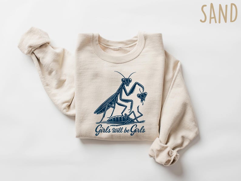 May include: Sand-colored sweatshirt with a navy blue graphic of a praying mantis and the text "Girls will be Girls." The design features a stylized illustration of a mantis and another insect. The sweatshirt has long sleeves.