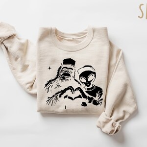 Puede incluir: Una sudadera beige con un estampado gr&aacute;fico negro de un Bigfoot y un extraterrestre haciendo un coraz&oacute;n con sus manos. El texto "SAND" est&aacute; impreso en la esquina superior derecha de la imagen.