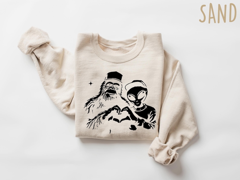 Puede incluir: Una sudadera beige con un estampado gr&aacute;fico negro de un Bigfoot y un extraterrestre haciendo un coraz&oacute;n con sus manos. El texto "SAND" est&aacute; impreso en la esquina superior derecha de la imagen.