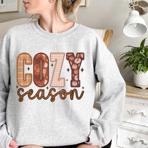 May include: A gray sweatshirt with the text "Cozy Season" in brown, orange, and white letters. The letters are decorated with plaid, floral, and pumpkin patterns.