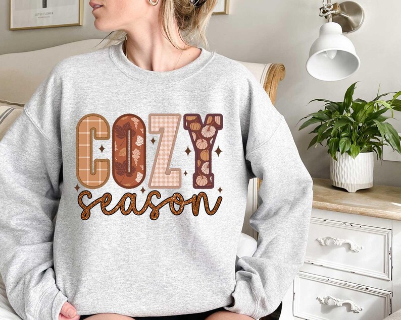 May include: A gray sweatshirt with the text "Cozy Season" in brown, orange, and white letters. The letters are decorated with plaid, floral, and pumpkin patterns.
