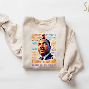 K&ouml;nnte beinhalten: Ein sandfarbenes Sweatshirt mit einem Portr&auml;t von Martin Luther King Jr. und dem Schriftzug "I Have a Dream". Das Design enth&auml;lt S&auml;tze wie "Gleichheit", "Friedlicher Widerstand" und "B&uuml;rgerrechte".