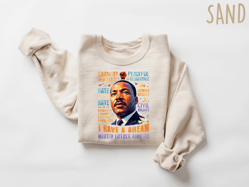 K&ouml;nnte beinhalten: Ein sandfarbenes Sweatshirt mit einem Portr&auml;t von Martin Luther King Jr. und dem Schriftzug "I Have a Dream". Das Design enth&auml;lt S&auml;tze wie "Gleichheit", "Friedlicher Widerstand" und "B&uuml;rgerrechte".