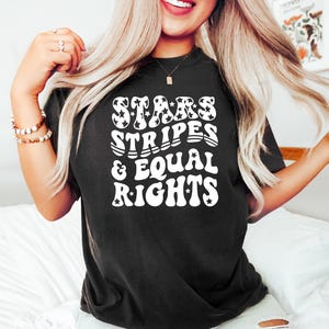 May include: Black t-shirt with the text "STARS STRIPES & EQUAL RIGHTS" in white, retro-style lettering. The word "STARS" features star-shaped designs. The shirt is a casual, comfortable style.
