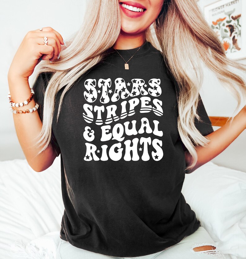May include: Black t-shirt with the text "STARS STRIPES & EQUAL RIGHTS" in white, retro-style lettering. The word "STARS" features star-shaped designs. The shirt is a casual, comfortable style.