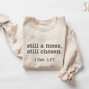 K&ouml;nnte beinhalten: Ein sandfarbenes Sweatshirt mit dem Text "still a mess, still chosen." und dem Bibelvers 1 Kor. 1:27 in Schwarz gedruckt. Die langen &Auml;rmel sind hochgekrempelt. Das Wort "SAND" steht in der oberen rechten Ecke.
