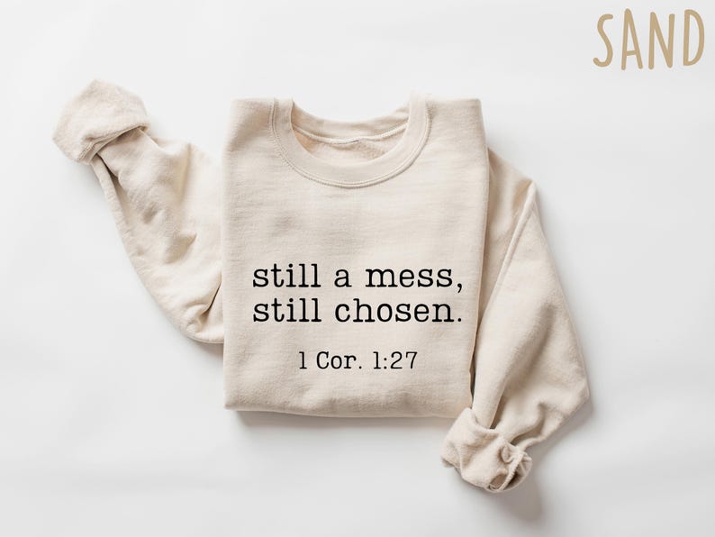 K&ouml;nnte beinhalten: Ein sandfarbenes Sweatshirt mit dem Text "still a mess, still chosen." und dem Bibelvers 1 Kor. 1:27 in Schwarz gedruckt. Die langen &Auml;rmel sind hochgekrempelt. Das Wort "SAND" steht in der oberen rechten Ecke.