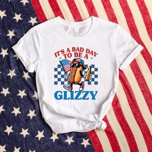 May include: White t-shirt with a cartoon hot dog wearing a red, white, and blue baseball cap and holding a beer. The text on the shirt reads "It's a bad day to be a Glizzy".