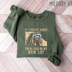 May include: Military green sweatshirt with a raccoon graphic. The graphic includes the text "IF I EVER GET RABIES YOU'RE HIGH ON MY BITIN' LIST". The sweatshirt is folded on a light wood grain surface.