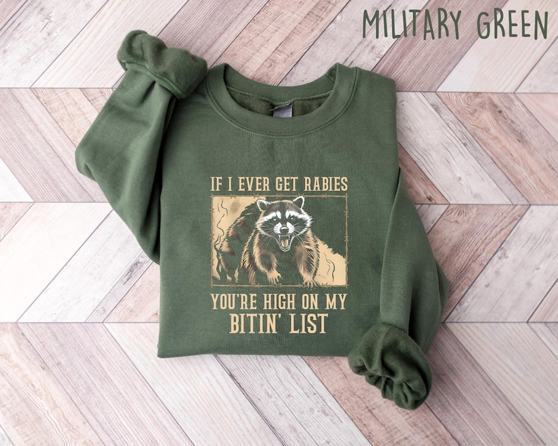 May include: Military green sweatshirt with a raccoon graphic. The graphic includes the text "IF I EVER GET RABIES YOU'RE HIGH ON MY BITIN' LIST". The sweatshirt is folded on a light wood grain surface.
