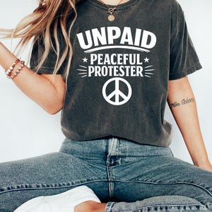 May include: Dark gray t-shirt with the words "UNPAID PEACEFUL PROTESTER" in white, along with a peace sign. The shirt is paired with blue jeans and white sneakers.