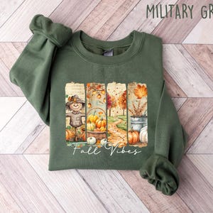May include: Military green sweatshirt with a fall-themed graphic. The graphic features a scarecrow, pumpkins, and the text "Fall Vibes". The sweatshirt is made of a soft material and is perfect for the autumn season.