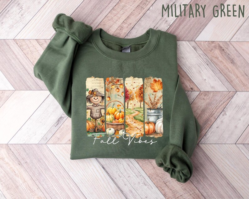 May include: Military green sweatshirt with a fall-themed graphic. The graphic features a scarecrow, pumpkins, and the text "Fall Vibes". The sweatshirt is made of a soft material and is perfect for the autumn season.