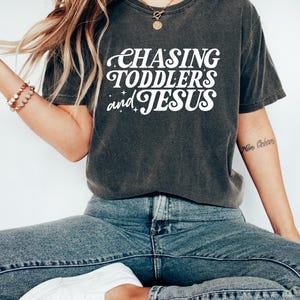 May include: Dark gray t-shirt with the text "CHASING TODDLERS and JESUS" in white, stylized lettering. The shirt is paired with blue jeans and white sneakers. The overall aesthetic is casual and comfortable.