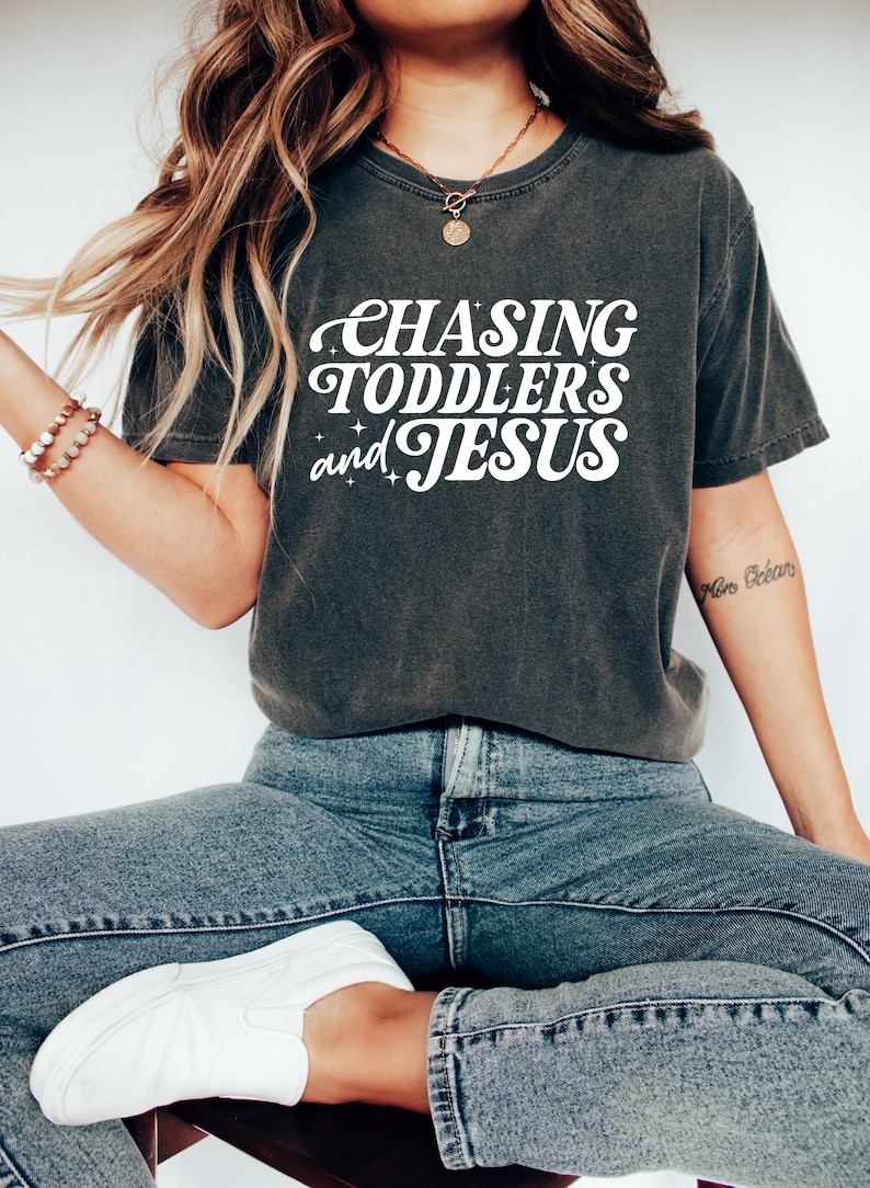 May include: Dark gray t-shirt with the text "CHASING TODDLERS and JESUS" in white, stylized lettering. The shirt is paired with blue jeans and white sneakers. The overall aesthetic is casual and comfortable.