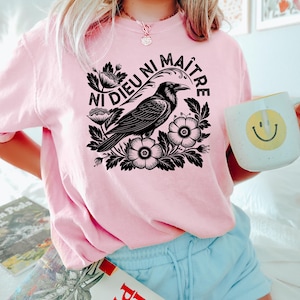 Op de afbeelding: Roze sweatshirt met een zwarte afbeelding van een raaf, bloemen en de tekst "Ni Dieu Ni Ma&icirc;tre". De persoon houdt een witte mok met een gele smiley vast. De persoon draagt een blauwe short.