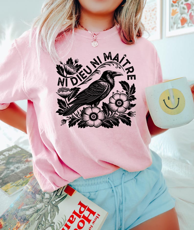 Op de afbeelding: Roze sweatshirt met een zwarte afbeelding van een raaf, bloemen en de tekst "Ni Dieu Ni Ma&icirc;tre". De persoon houdt een witte mok met een gele smiley vast. De persoon draagt een blauwe short.