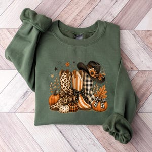 May include: An olive green crewneck sweatshirt featuring a graphic print of three cowboy boots, a cowboy hat, pumpkins, and autumn leaves. The boots display leopard print, vertical stripes, and buffalo plaid patterns with black bows.