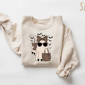 May include: A sand-colored sweatshirt featuring a ghost graphic. The ghost wears sunglasses, a leopard print bow, and carries a leopard print bag and a coffee cup. Black bats and stars are also part of the design.