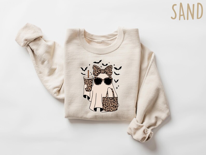 May include: A sand-colored sweatshirt featuring a ghost graphic. The ghost wears sunglasses, a leopard print bow, and carries a leopard print bag and a coffee cup. Black bats and stars are also part of the design.