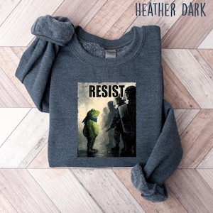 May include: Heather dark gray sweatshirt featuring a graphic of a frog facing a line of silhouettes with the word "RESIST". The frog is wearing a blue scarf. The sweatshirt is a dark gray color.