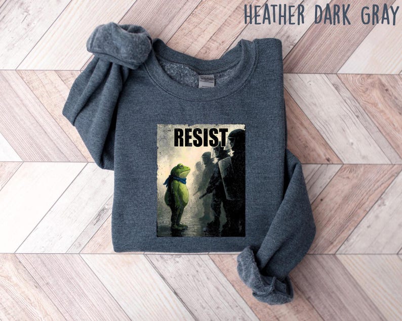 May include: Heather dark gray sweatshirt featuring a graphic of a frog facing a line of silhouettes with the word "RESIST". The frog is wearing a blue scarf. The sweatshirt is a dark gray color.