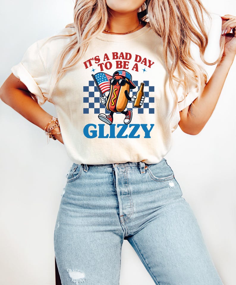 May include: A white t-shirt with a graphic of a hot dog wearing a baseball cap and sunglasses holding a flag and a beer. The text on the shirt reads "It's a bad day to be a Glizzy".