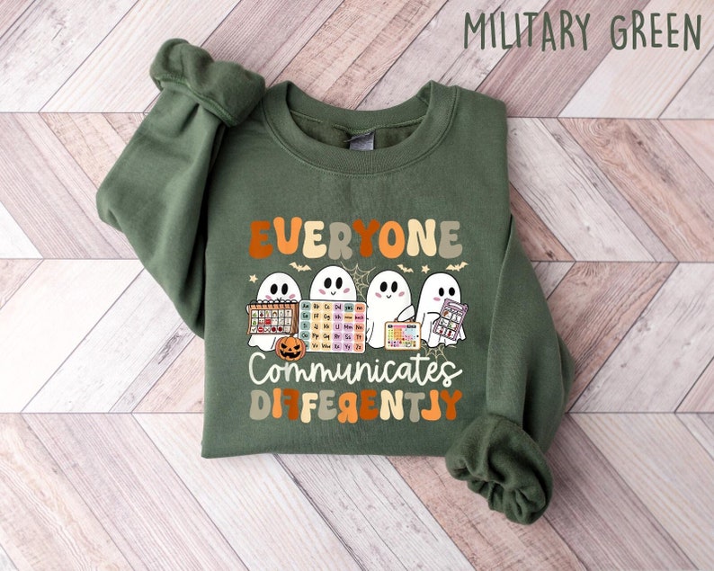 Może przedstawiać: Zielona bluza w kolorze wojskowej zieleni z napisem "EVERYONE COMMUNICATES DIFFERENTLY" i ilustracjami duch&oacute;w trzymających przybory szkolne. Projekt zawiera dynię, pajęczynę i układ okresowy. Bluza wykonana jest z miękkiego materiału.