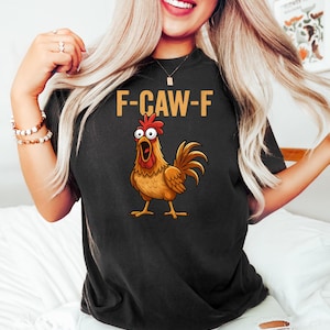Puede incluir: Camiseta negra con el texto "F-CAW-F" y una ilustraci&oacute;n de un gallo de dibujos animados. El gallo es marr&oacute;n, con cresta roja y pico abierto.
