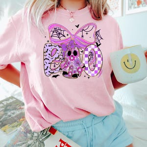 May include: A light pink t-shirt with a Halloween-themed graphic. The graphic spells "BOO" with a floral ghost and a purple bow with spiderwebs. The shirt is paired with light blue shorts. A mug with a smiley face is held in the hand.
