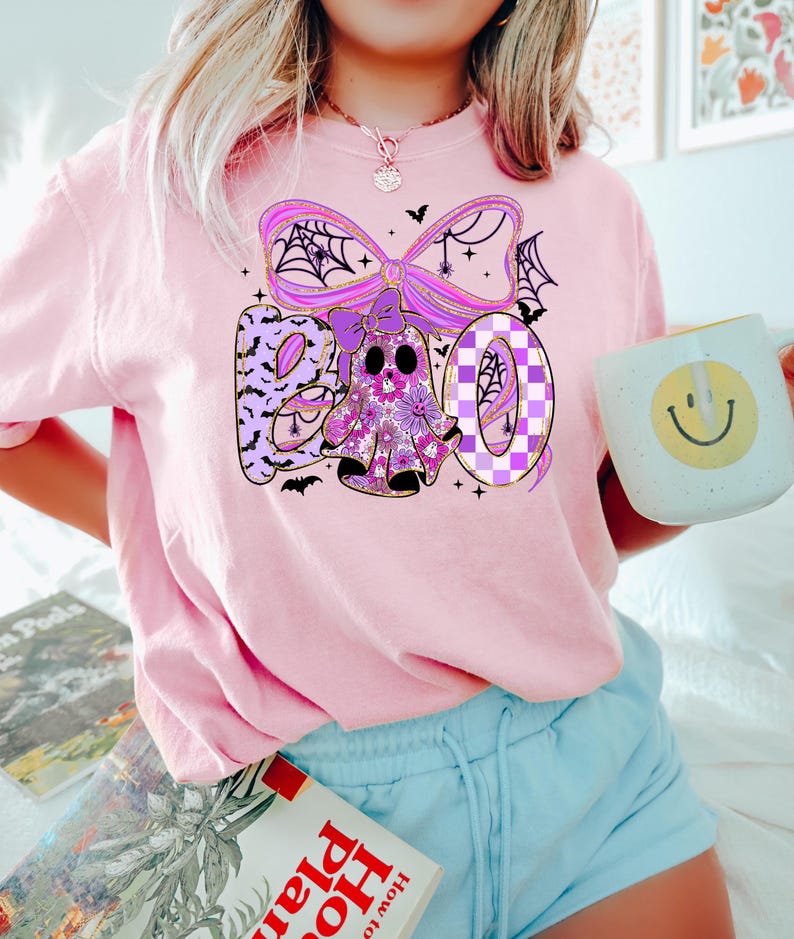 May include: A light pink t-shirt with a Halloween-themed graphic. The graphic spells "BOO" with a floral ghost and a purple bow with spiderwebs. The shirt is paired with light blue shorts. A mug with a smiley face is held in the hand.