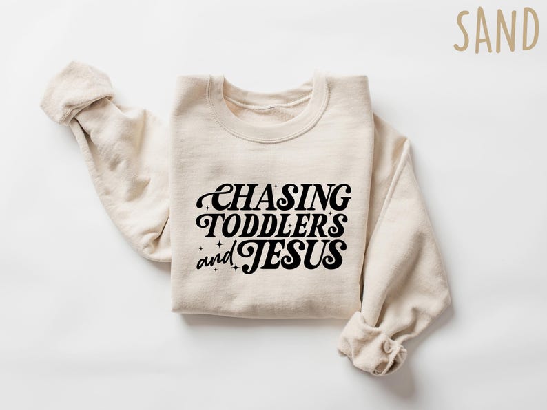 May include: A sand-colored sweatshirt with the phrase "CHASING TODDLERS and JESUS" in black script. The sweatshirt is folded with the sleeves rolled up, and the word "SAND" is in the upper right corner.