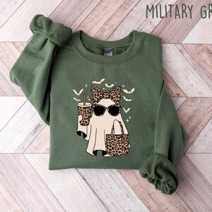 May include: Military green sweatshirt featuring a ghost graphic with sunglasses, a bow, and a leopard print cup and purse. The design includes bats and stars. The text "MILITARY GREEN" is at the top.
