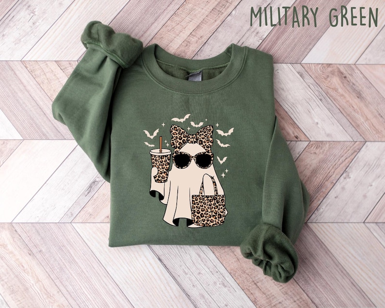 May include: Military green sweatshirt featuring a ghost graphic with sunglasses, a bow, and a leopard print cup and purse. The design includes bats and stars. The text "MILITARY GREEN" is at the top.
