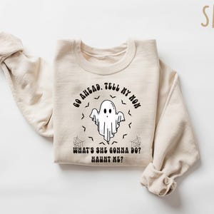 May include: A sand-coloured sweatshirt featuring the text "GO AHEAD. TELL MY MOM WHAT'S SHE GONNA DO? HAUNT ME?" and a ghost graphic. The design includes bats and spiderwebs, ideal for Halloween.