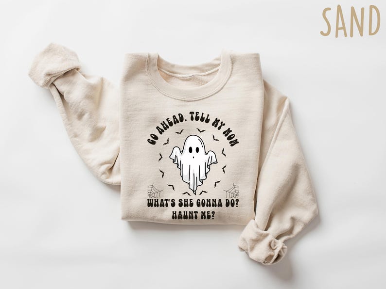 May include: A sand-coloured sweatshirt featuring the text "GO AHEAD. TELL MY MOM WHAT'S SHE GONNA DO? HAUNT ME?" and a ghost graphic. The design includes bats and spiderwebs, ideal for Halloween.