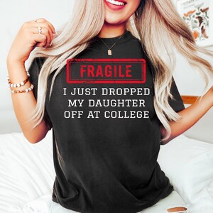 May include: Black t-shirt with the text "FRAGILE" in a red rectangle, and the text "I JUST DROPPED MY DAUGHTER OFF AT COLLEGE" in white. The shirt is worn by a person with long blonde hair.