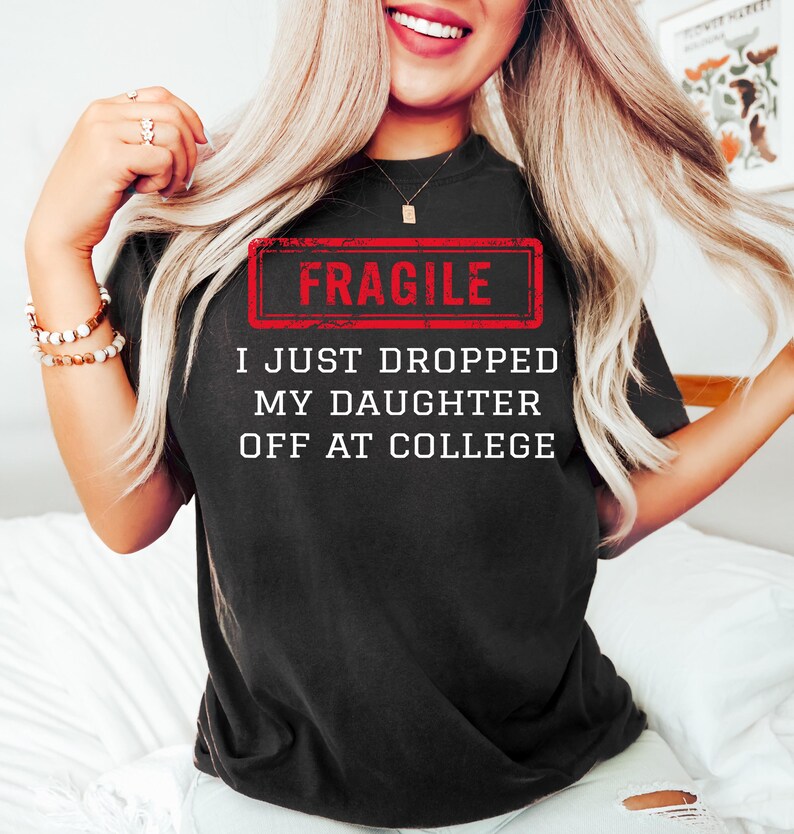 May include: Black t-shirt with the text "FRAGILE" in a red rectangle, and the text "I JUST DROPPED MY DAUGHTER OFF AT COLLEGE" in white. The shirt is worn by a person with long blonde hair.