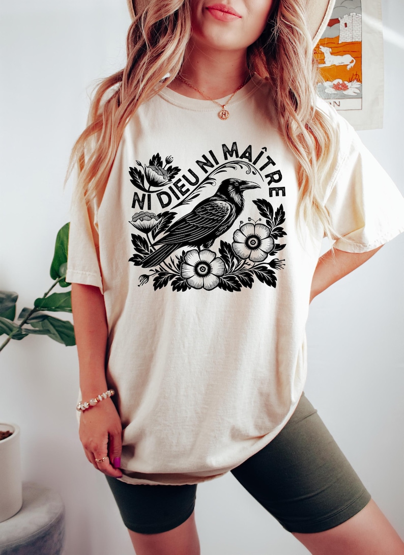 Op de afbeelding: Cr&egrave;mekleurig T-shirt met een zwarte afbeelding van een raaf omringd door bloemen, met de Franse tekst "Ni Dieu Ni Ma&icirc;tre" (Noch God noch Meester) in een cirkel. Het shirt heeft een relaxte pasvorm.