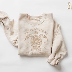 May include: A beige sweatshirt with a graphic of a sea turtle and the text "The Gulf of Mexico Est. 1550 Stay Salty and Resist".
