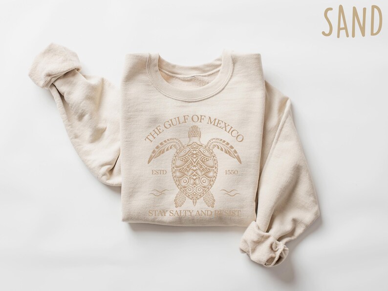 May include: A beige sweatshirt with a graphic of a sea turtle and the text "The Gulf of Mexico Est. 1550 Stay Salty and Resist".